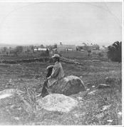 SA0070 - William Briggs sits on a rock in a field; buildings and stone walls are in the distance. Identified on the reverse. Modern photos derived from a stereograph., Winterthur Shaker Photograph and Post Card Collection 1851 to 1921c